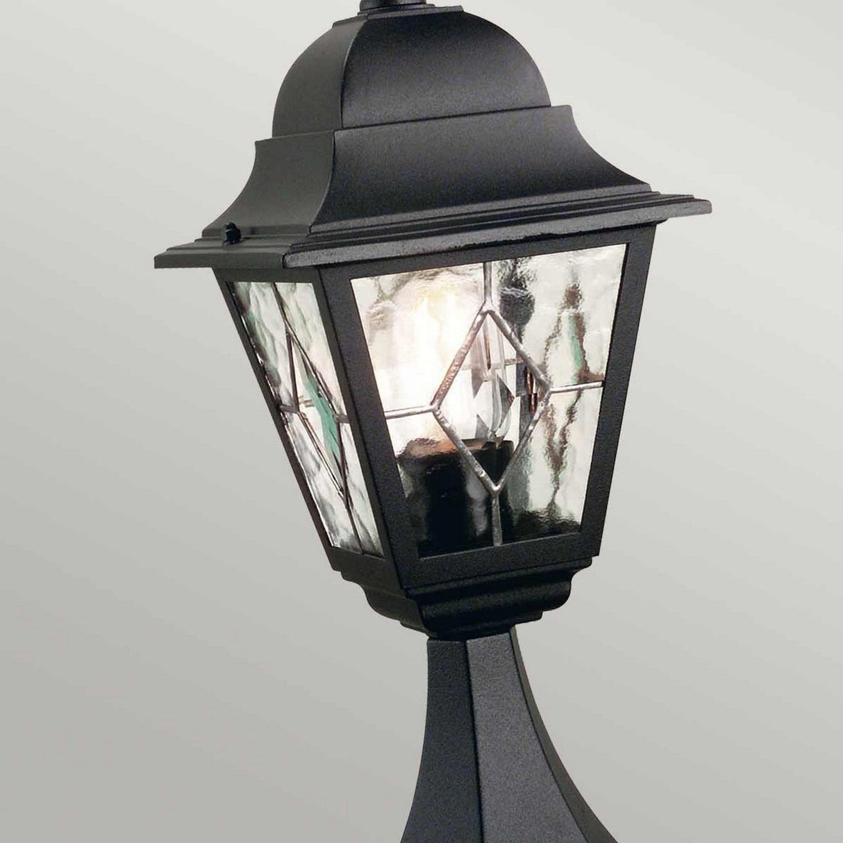 The Norfolk 1 Light Pedestal Light - Black is an outdoor lantern showcasing decorative design elements and hand-leaded glass panels. It's mounted on a post and seamlessly enhances any outdoor lighting setup by casting a gentle glow on a pale backdrop.