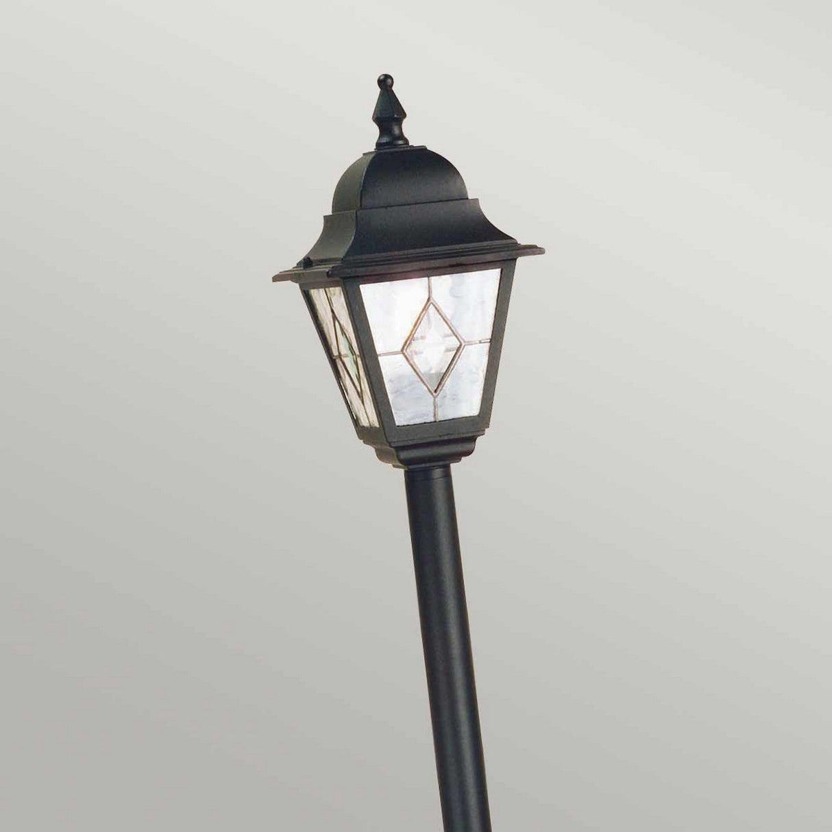 The Norfolk 1 Light Pillar Light in black, showcasing a traditional lantern-style top with hand-leaded glass panels and a diamond pattern, is displayed against a plain, light gray background.