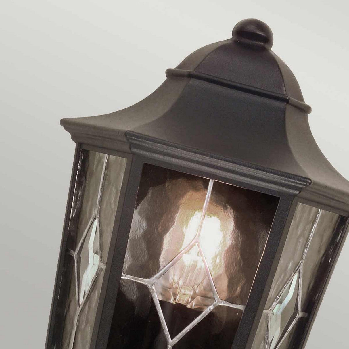 A close-up of the Norfolk 1 Light Outdoor Wall Light in black shows a lit bulb glowing inside. The hand-leaded glass panels, adorned with a decorative diamond pattern, emit a warm and inviting light against a pale backdrop, ideal for sophisticated lantern styling.