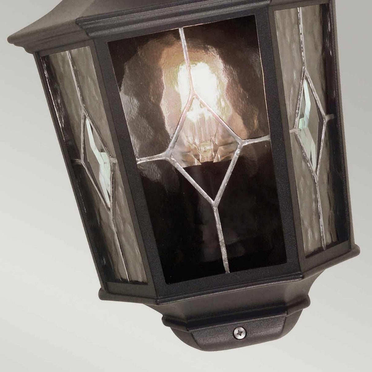 The Norfolk 1 Light Outdoor Wall Light in black features a decorative design with hand-leaded glass panels adorned with diamond-shaped accents. When illuminated, the light bulb inside casts a warm and inviting glow.