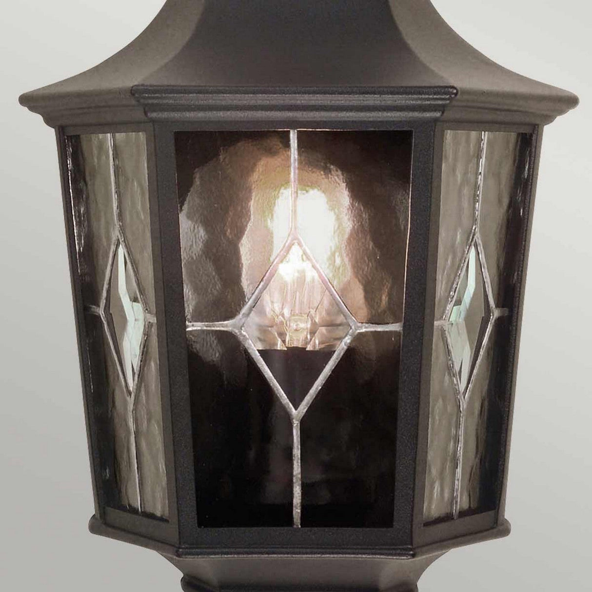 A close-up of the Norfolk 1 Light Outdoor Wall Light - Black showcases a lantern design with a glowing bulb inside. Its black metal frame complements glass panels featuring a diamond-shaped design, adding an elegant touch to your outdoor decor.