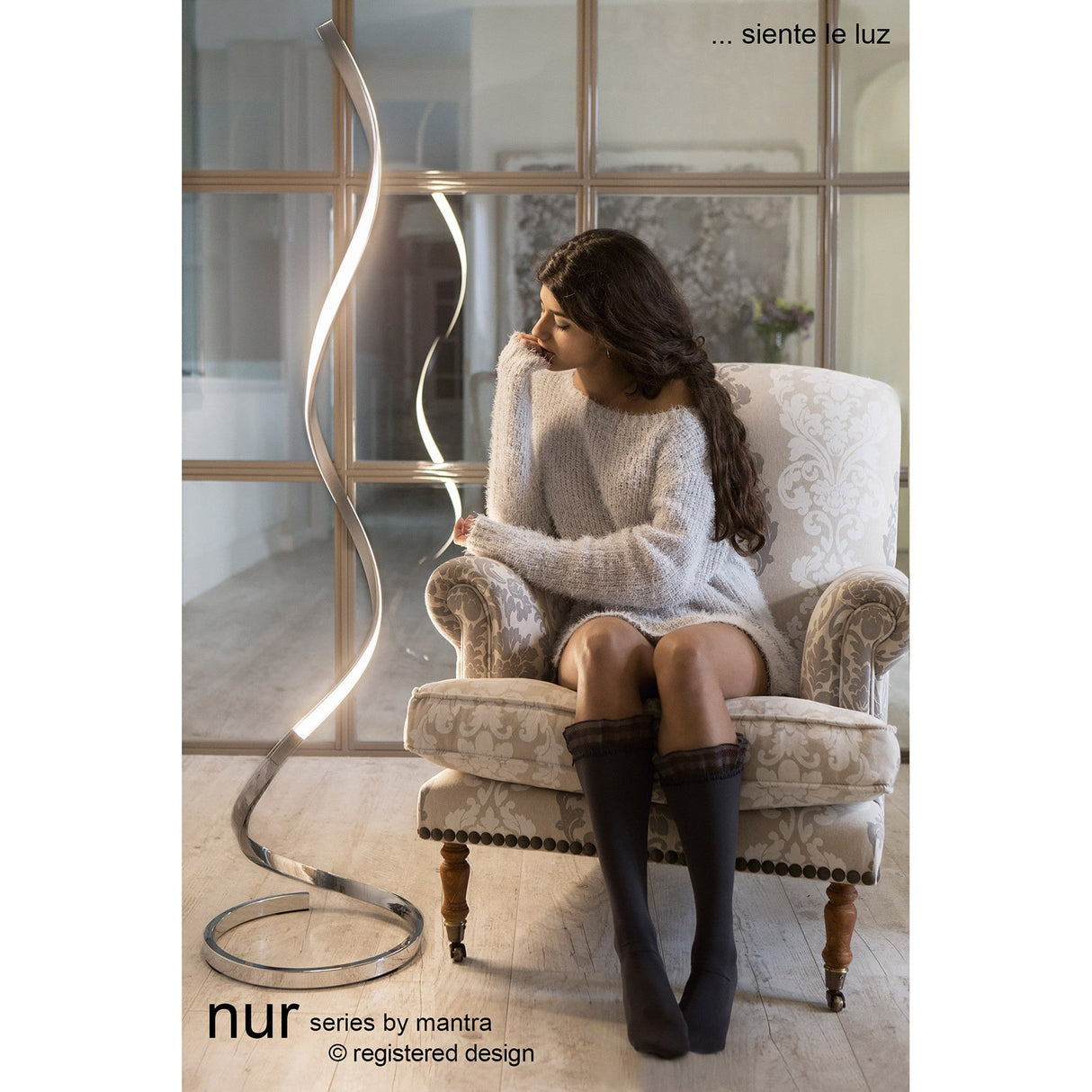 A woman in a cozy sweater and knee-high socks sits on an ornate armchair, gently touching the Nur Dimmable LED Floor Lamp 20W 3000K - Silver & Chrome, which emits soft ambient light. Large glass windows frame the background with the text: “nur series by mantra.”.
