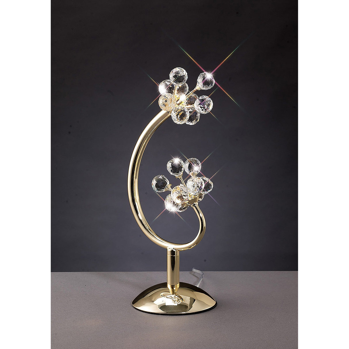 The Octavia 2 Light Crystal Table Lamp in French Gold, featuring a curved stem and clusters of clear crystal spheres like stylized flowers, is displayed on a gray surface against a dark background, providing elegant ambient lighting.