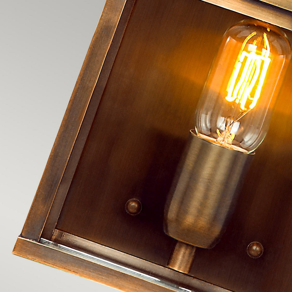 A close-up of the Old Bailey 1 Light Outdoor Wall Light in aged brass shows a vintage-style Edison bulb glowing warmly inside its rectangular metal housing. The background features a gradient from light to dark gray.