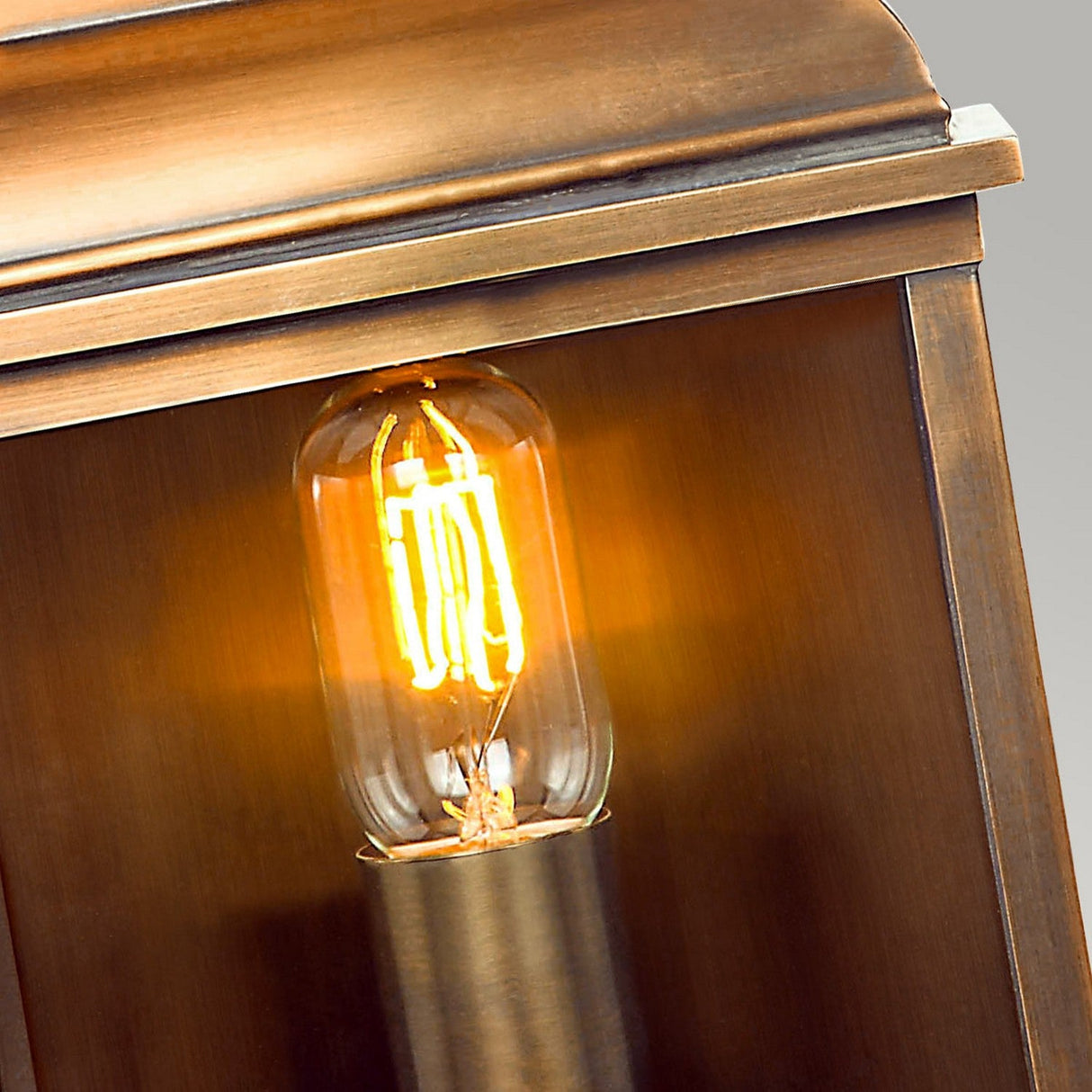 A close-up of the Old Bailey 1 Light Outdoor Wall Light, featuring a vintage-style Edison bulb that emits a warm orange glow. It is housed in a fixture with an aged brass finish and a rectangular shape. The neutral gray background highlights the warm tone of the light beautifully.