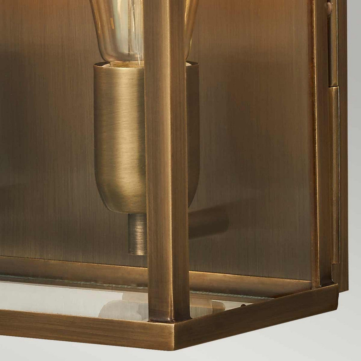 Close-up of the Old Bailey 2 Light Outdoor Wall Light in aged brass, highlighting its vintage-style bronze and glass design with a single bulb in a metallic holder. The solid brass fixture showcases a geometric pattern with sharp angles and an antique finish, casting a warm, golden glow.