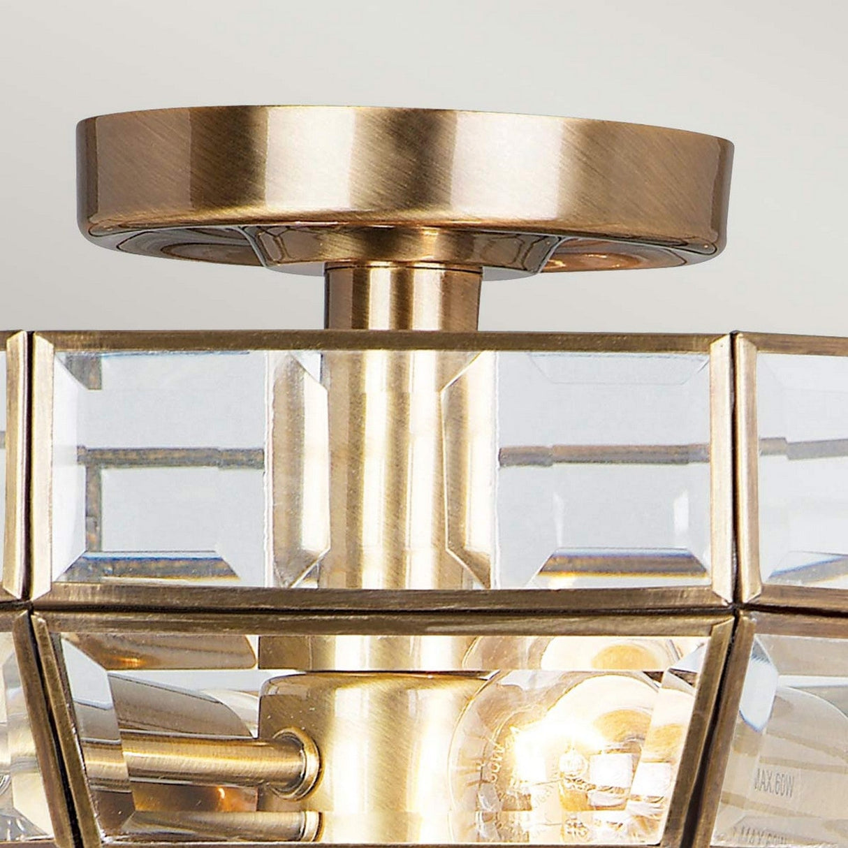 Close-up of the Old Park 2 Light Semi Flush in antique brass. This fixture showcases a semi-flush design with a metallic base and clear, angular glass panes surrounding the bulbs, casting a warm glow through its geometric patterns.