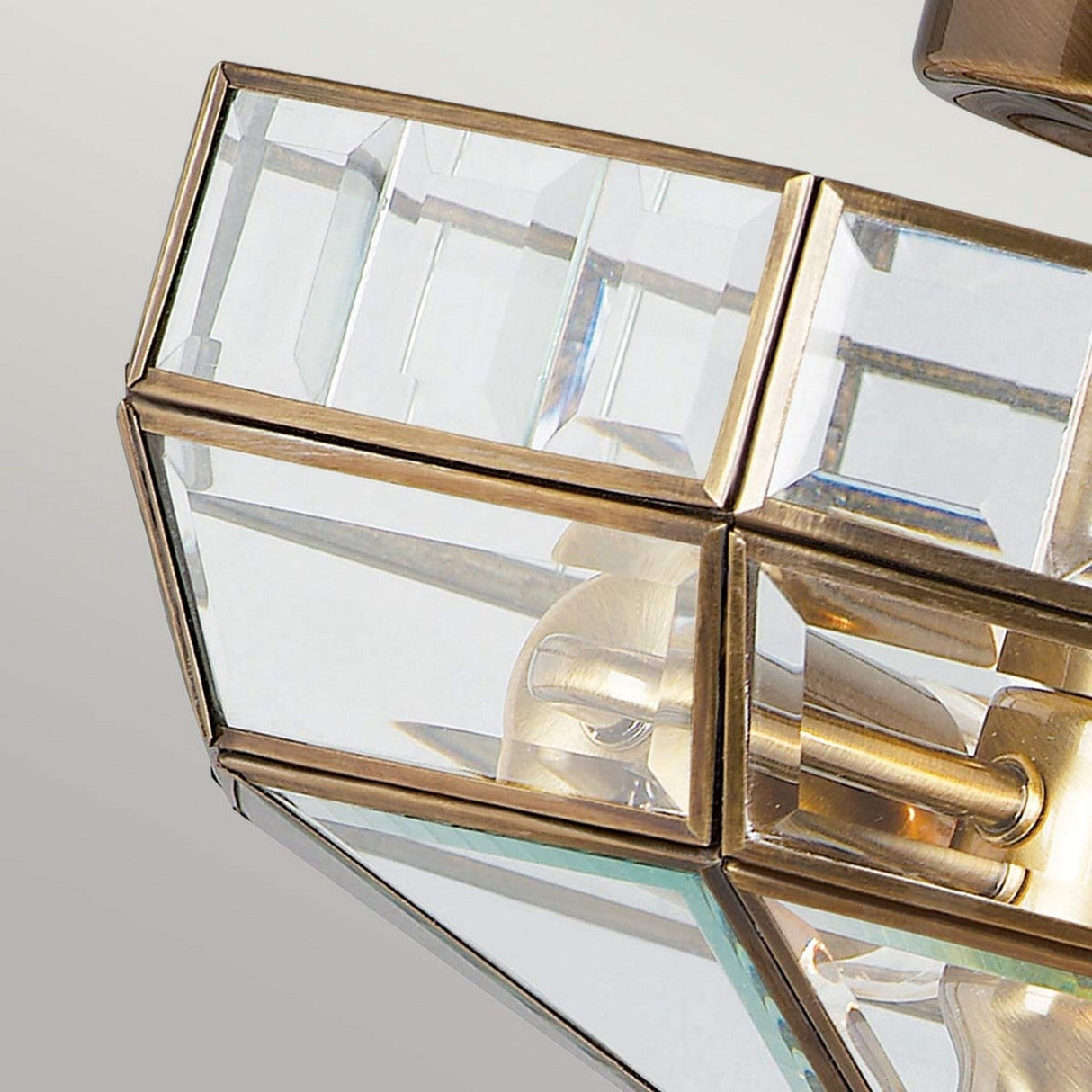 A close-up view of the Old Park 2 Light Semi Flush in Antique Brass reveals its geometric pendant design with an antique brass frame and clear glass panels. This fixture beautifully reflects light and highlights its precise angles, exemplifying the craftsmanship and modern aesthetic founded on solid brass construction.