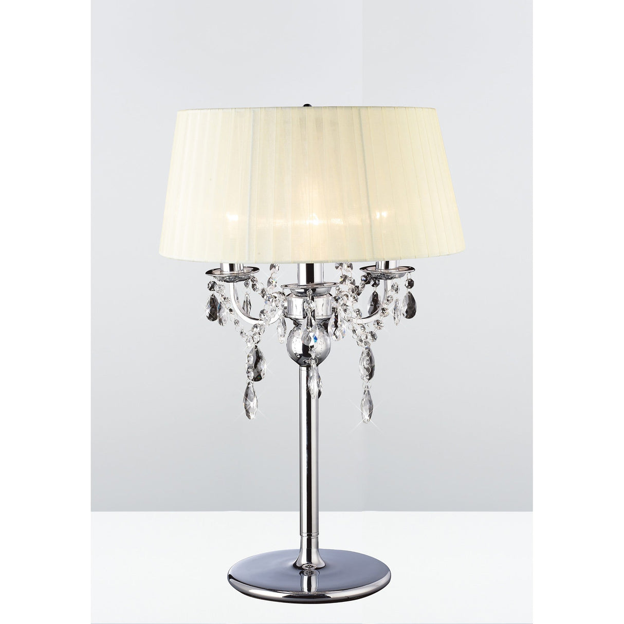 The Olivia 3 Light Crystal Table Lamp, featuring a chrome base, cream pleated shade, and crystal pendants, is displayed on a white surface with a light background.