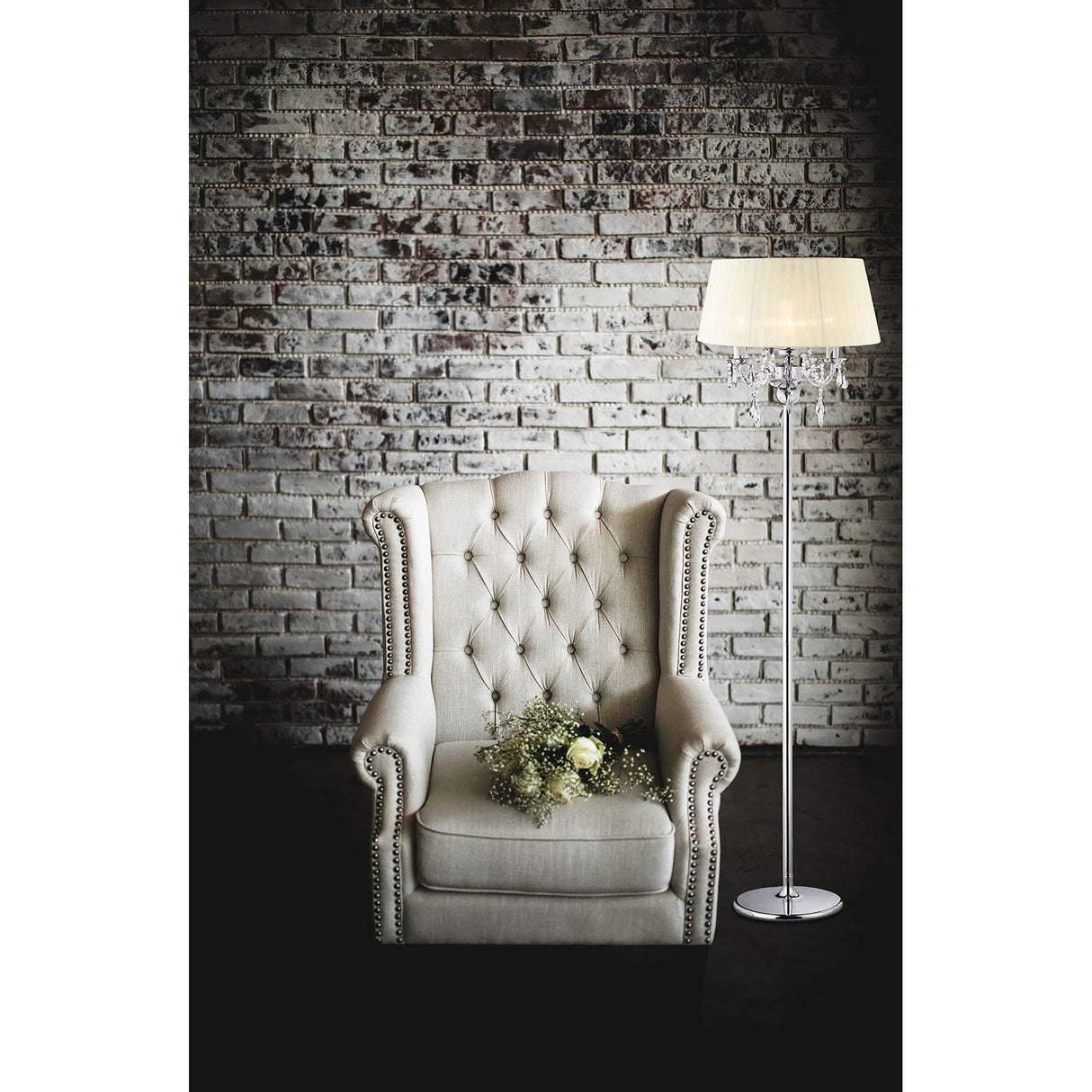 A tufted beige armchair with white flowers sits beside the Olivia 3 Light Crystal Floor Lamp—chrome with a cream shade—set against a textured white brick wall.