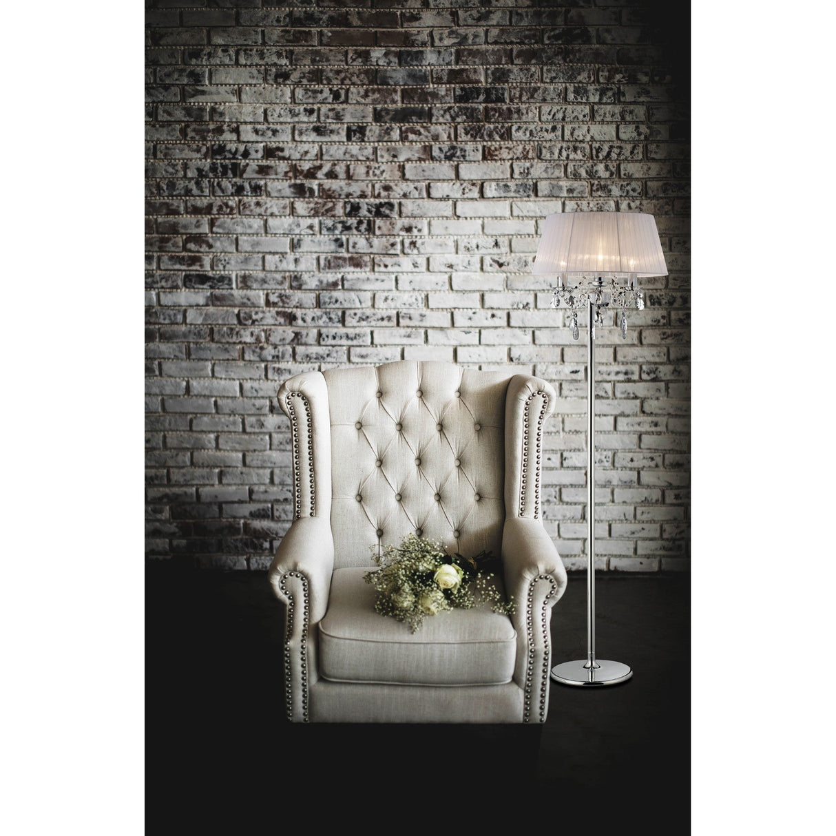 A beige tufted armchair with a bouquet of flowers sits in front of a white brick wall, next to the Olivia 3 Light Crystal Floor Lamp in chrome with a white shade.