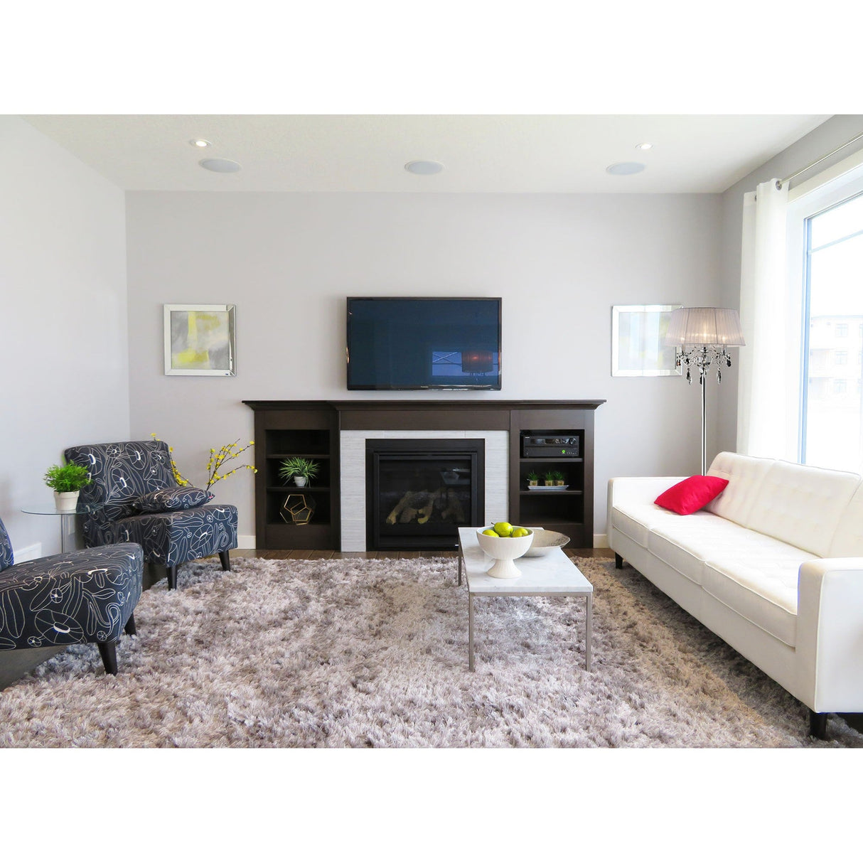 Modern living room with a white sofa, patterned armchairs, fireplace under a wall-mounted TV, shaggy gray rug, decor-topped coffee table, and large window lit by the Olivia 3 Light Crystal Floor Lamp - Chrome With White Shade.