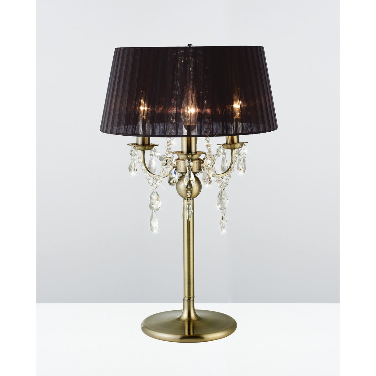 The Olivia 3 Light Crystal Table Lamp in brass features a round base, three candle-style lights, hanging crystal accents, and a semi-transparent pleated black shade, set against a plain light background.