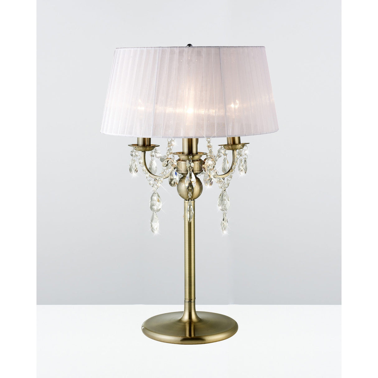 The Olivia 3 Light Crystal Table Lamp features an antique brass finish, white pleated shade, three candle-style bulbs, and hanging crystal ornaments—ideal for elegant interiors. Shown against a plain white background.