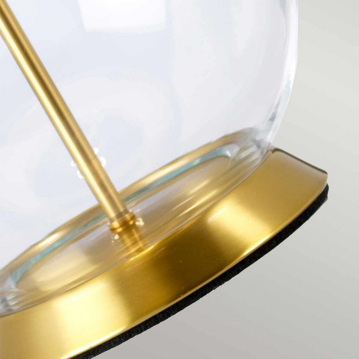 A close-up of the Orb 1 Light Table Lamp shows off its clear glass shade, paired with an aged brass base. A gold rod rises from the base to support the lamp, which is set against a plain background.