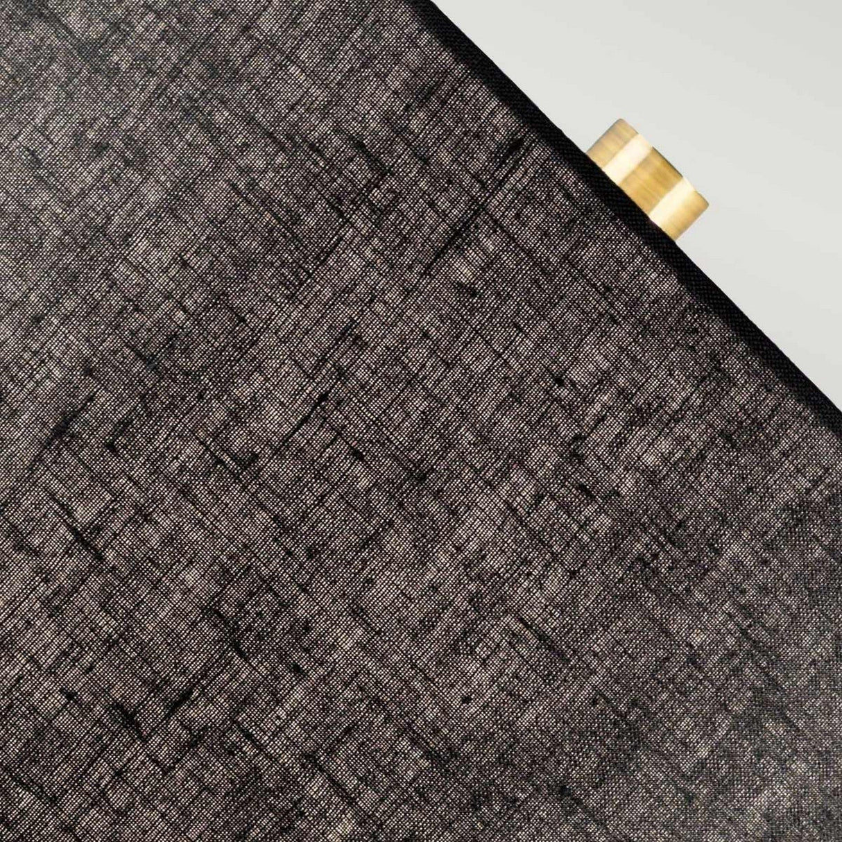 A close-up of the Orb 1 Light Table Lamp, featuring its black textured geometric shade partially revealing the aged brass cylindrical base on the top right corner against a light background.