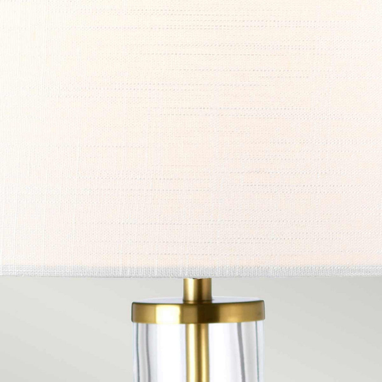 Close-up of the Orb 1 Light Table Lamp characterized by a white drum shade and an aged brass-accented base crafted from clear acrylic. The soft gray background highlights its minimalist and elegant design, making it an ideal addition to any space.