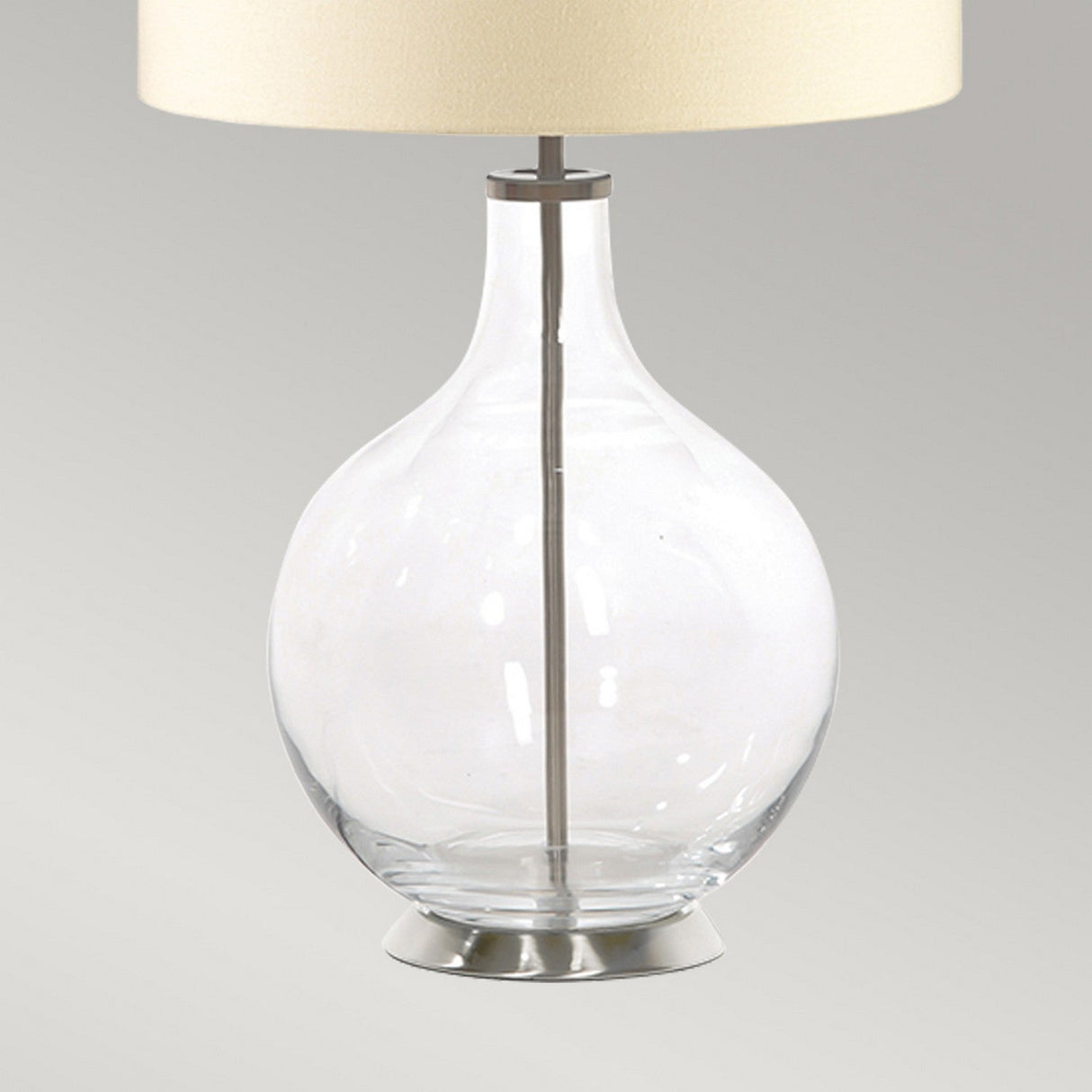 The Orb 1 Light Table Lamp boasts a clear glass base and a beige shade, gracefully situated on a metallic circular platform against a plain, light gray background.