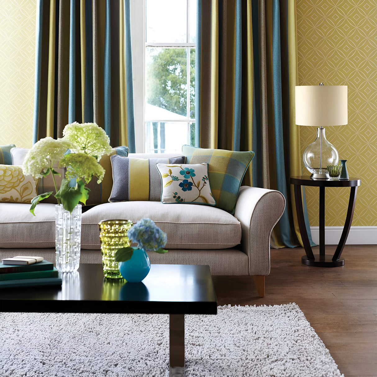 A cozy living room featuring a beige sofa with floral and checkered cushions. A dark wood coffee table is adorned with vases of flowers. Light streams through a window framed by blue and brown curtains, next to a round side table topped with the Orb 1 Light Table Lamp that has a clear glass base and beige shade.