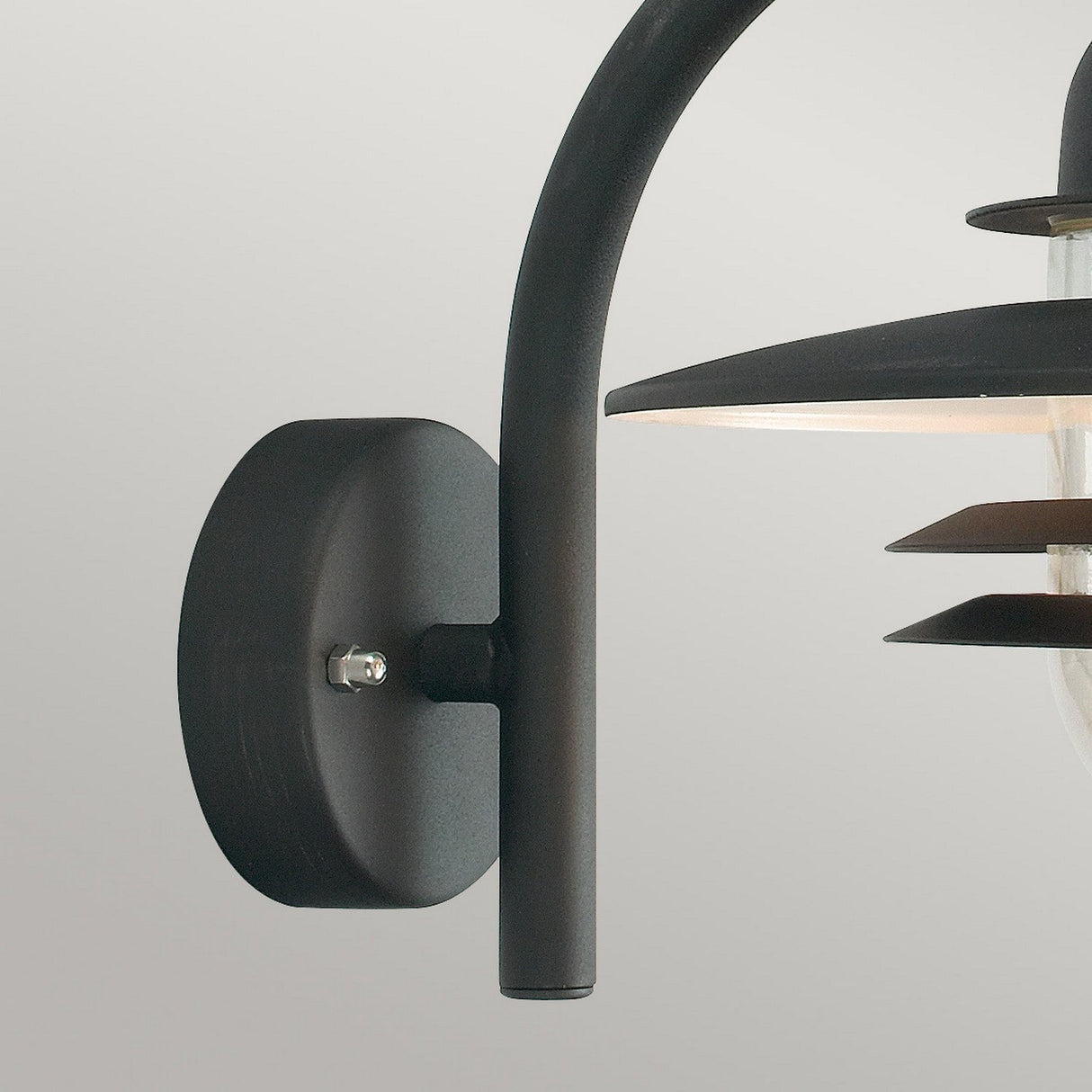 Close-up of the Oslo Outdoor 1 Light Wall Lantern in black, showcasing its modern design with a cylindrical base and curved arm. Reminiscent of Scandinavian aesthetics, this elegant fixture is adorned with layered horizontal shades and is mounted on a light gray wall, harmoniously combining function with style in outdoor lighting.