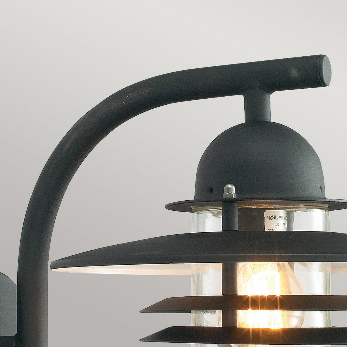 Close-up of the Oslo Outdoor 1 Light Wall Lantern in black, highlighting its Scandinavian design with a modern aesthetic. It boasts a curved arm and concentric circular shades, featuring a visible glowing bulb encased in a clear glass cover.