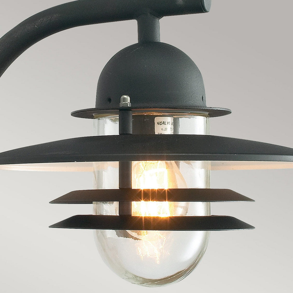 A close-up showcases the Oslo Outdoor 1 Light Wall Lantern - Black, featuring a Scandinavian design with a transparent glass shade and black metal accents. The illuminated bulb inside accentuates the elegant design of the wall lantern against a plain backdrop.