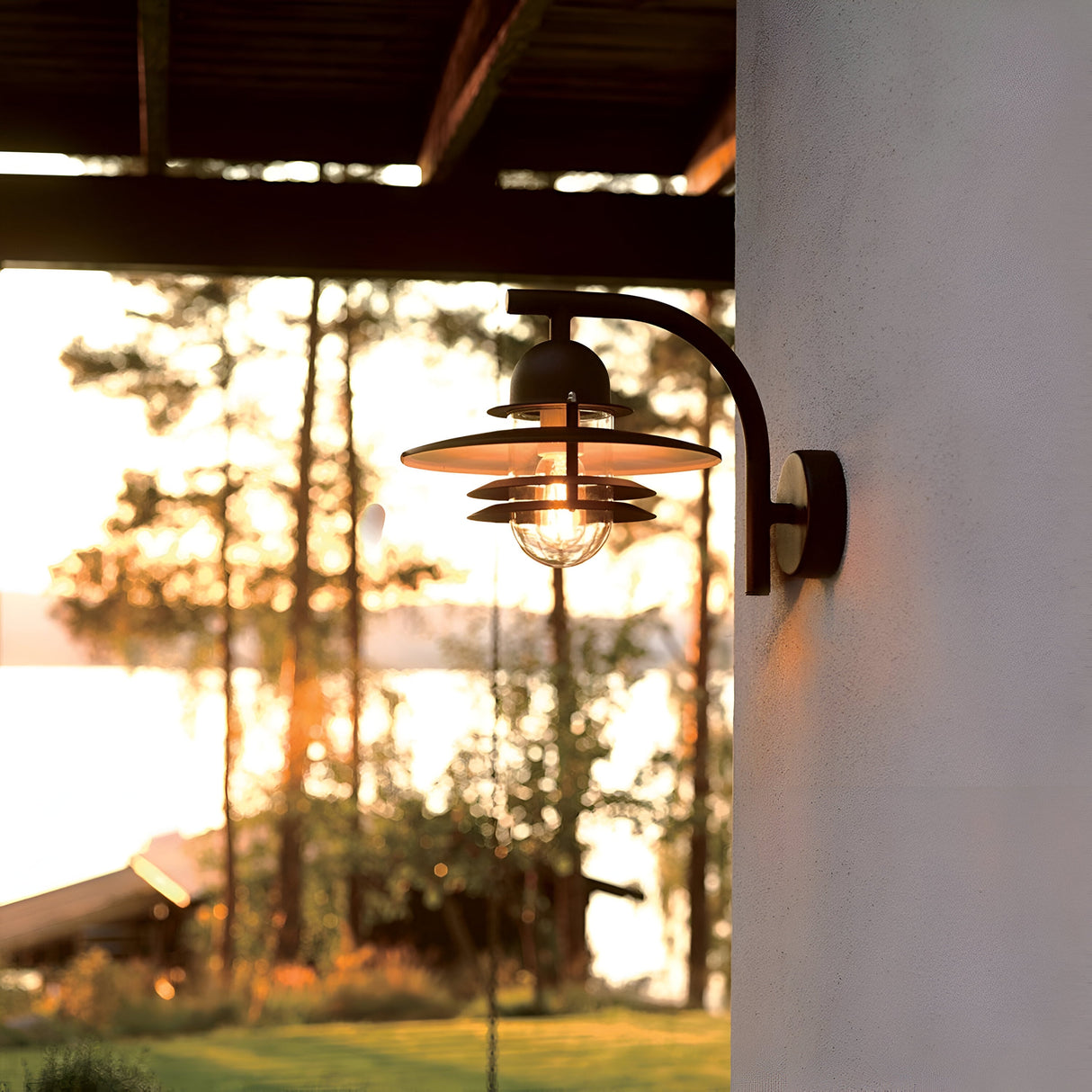 The Oslo Outdoor 1 Light Wall Lantern, with its vintage design, is mounted on a white wall, casting a warm glow as the sun sets. In the background lies a picturesque scene of trees, a tranquil lake, and a small wooden structure illuminated in golden light—an ideal representation of Scandinavian outdoor lighting.