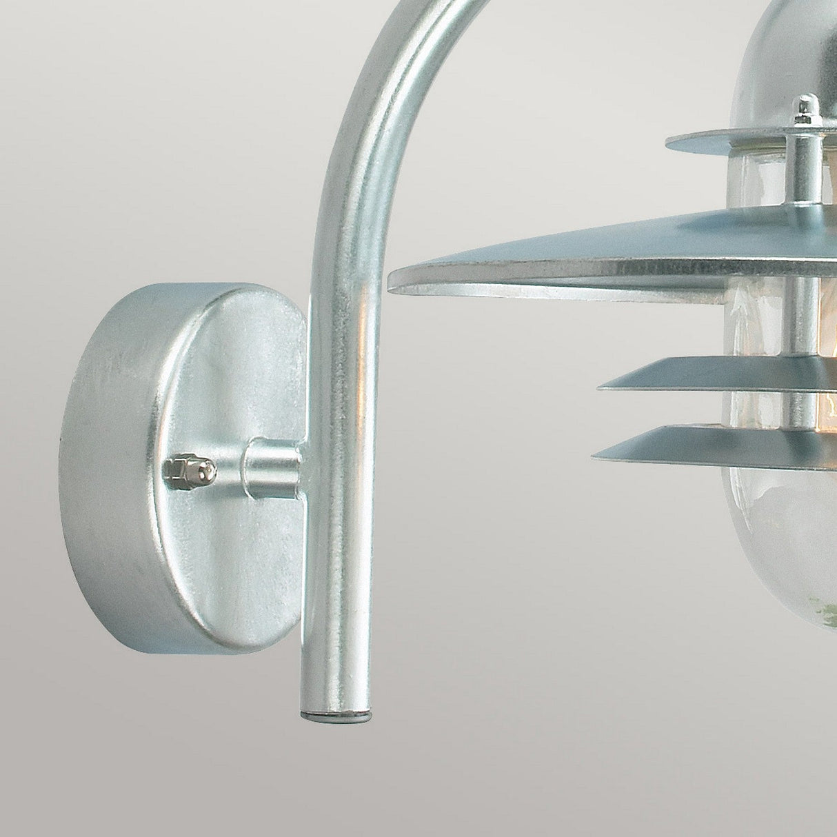 Close-up of the Oslo Outdoor 1 Light Wall Lantern - Silver; a contemporary metal wall sconce made from galvanized steel, featuring a cylindrical base and an elegantly arched arm. It includes layered circular plates and a clear glass cover, all set against a neutral background to highlight its IP55-rated minimalist design.