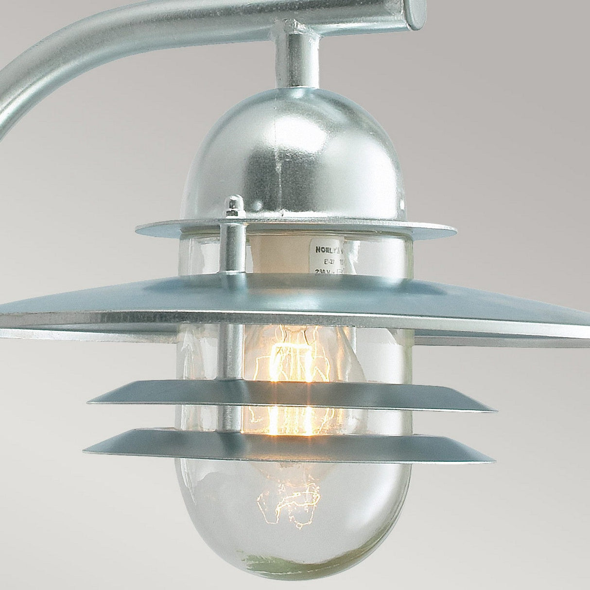 The Oslo Outdoor 1 Light Wall Lantern - Silver showcases a contemporary design with a clear, oval-shaped glass cover. It emits a warm glow from inside and is crafted from galvanized steel with an IP55 rating. Its industrial style is highlighted by several horizontal metal bands, creating a sleek appearance.