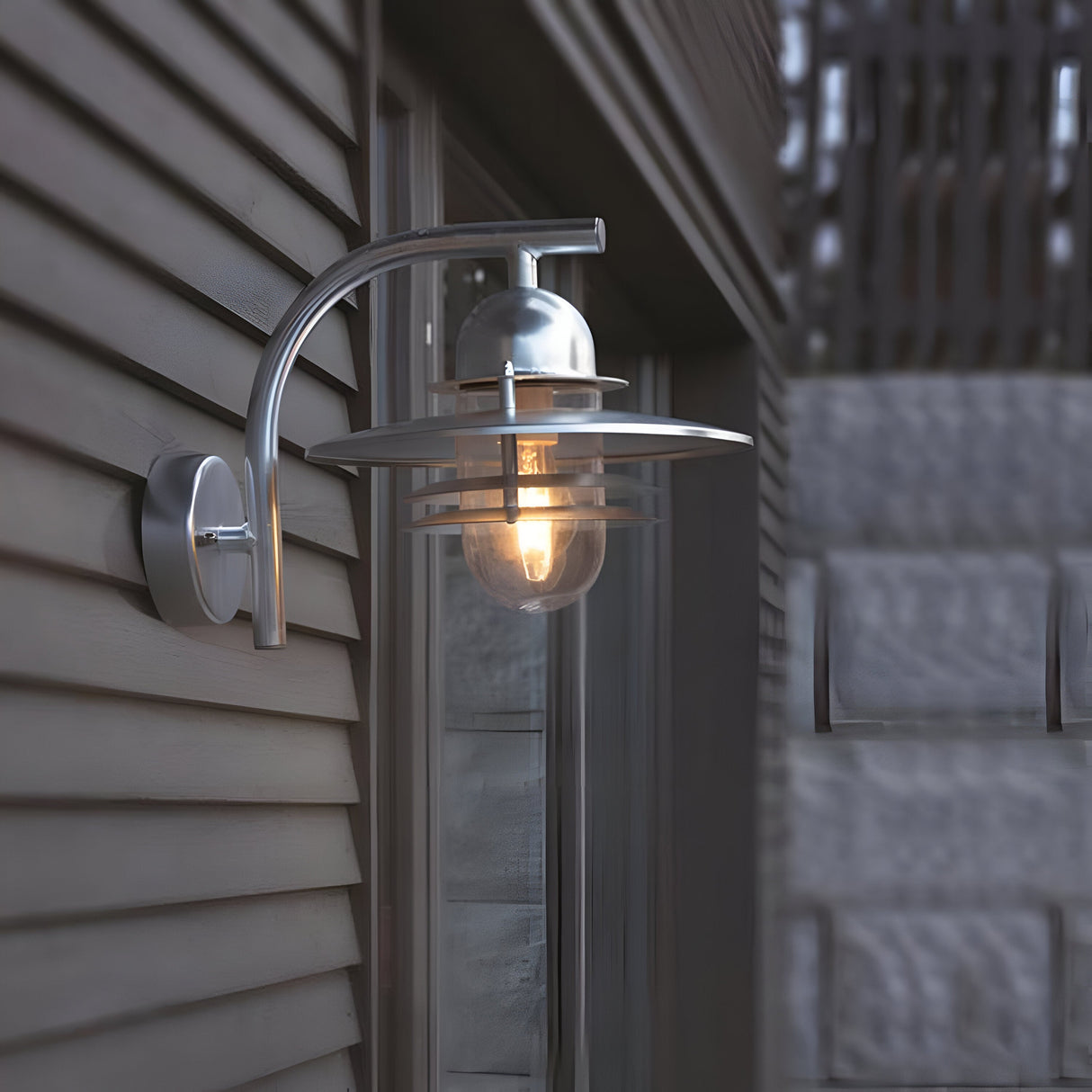 The Oslo Outdoor 1 Light Wall Lantern - Silver, made from galvanized steel, showcases a stylish curved metal arm and a transparent glass bulb cover. Installed on the exterior wall of a home, this IP55-rated lantern emits a warm glow across the siding and its surroundings.