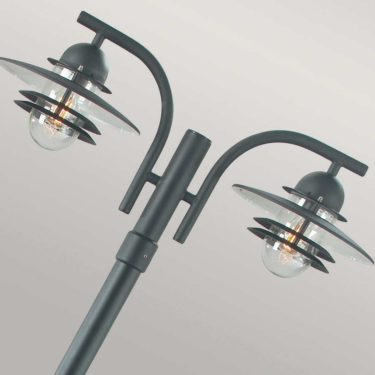 Introducing the Oslo 2 Light Lamp Post - Black, a modern exterior lighting piece. It boasts two curved arms and round glass enclosures with a visible light bulb. The black metal finish contrasts strikingly against a neutral gray background, reminiscent of stylish galvanized steel designs.