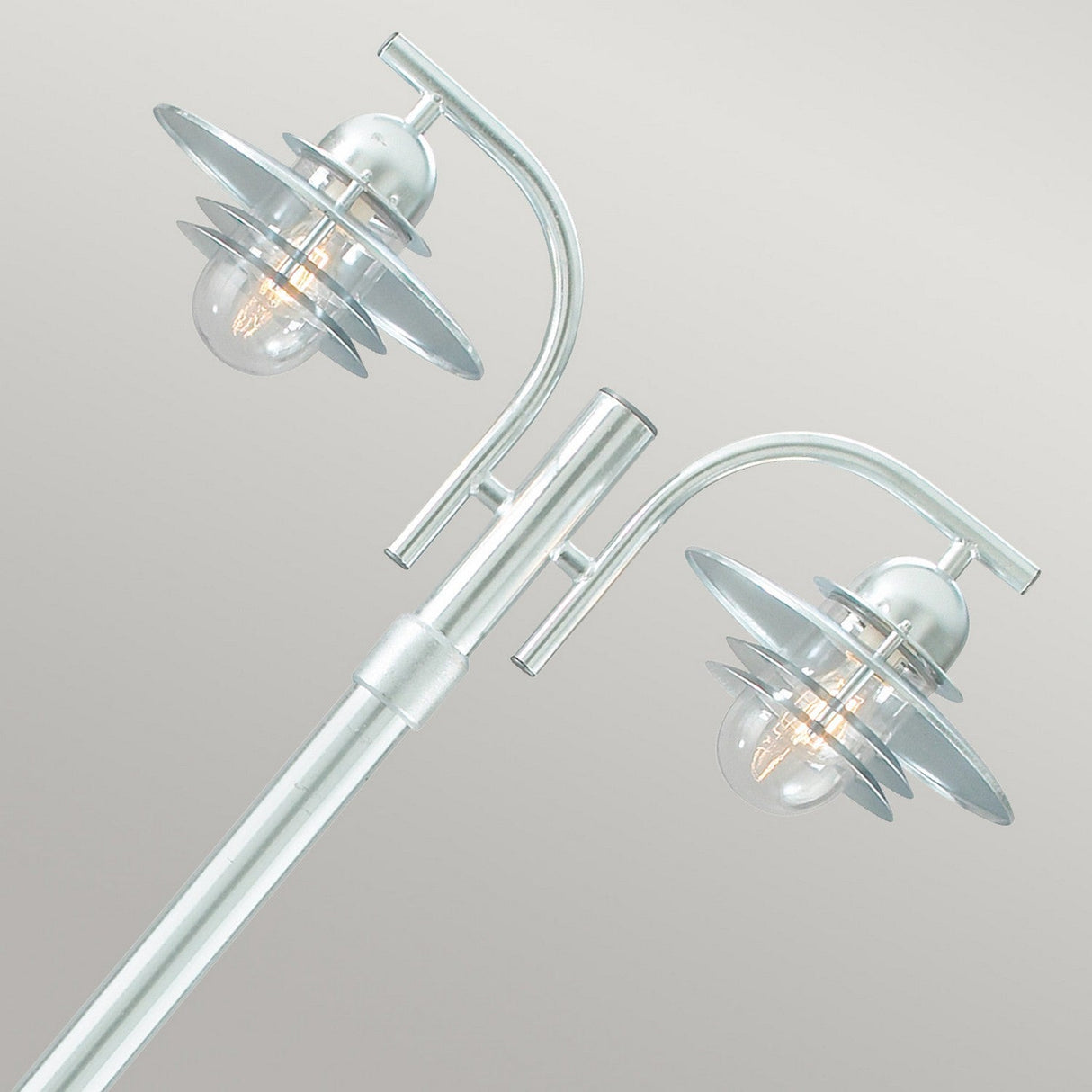 The Oslo 2 Light Lamp Post - Silver is a contemporary outdoor lighting fixture with a sleek metallic finish, made from galvanized steel with an IP55 rating. It features two bulbs housed in circular metal shades atop a silver pole, casting a warm glow against its neutral surroundings.
