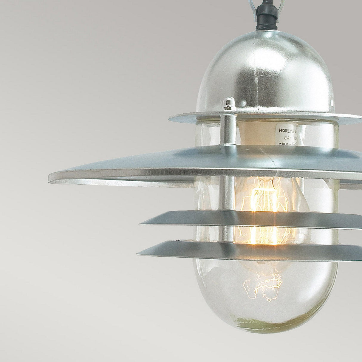 Introducing the Oslo Outdoor 1 Light Chain Pendant in Silver: This light fixture boasts a sleek Scandinavian design, crafted from galvanized steel with round metal layers. It features a clear glass bulb that emits a warm glow against a plain, light gray backdrop.