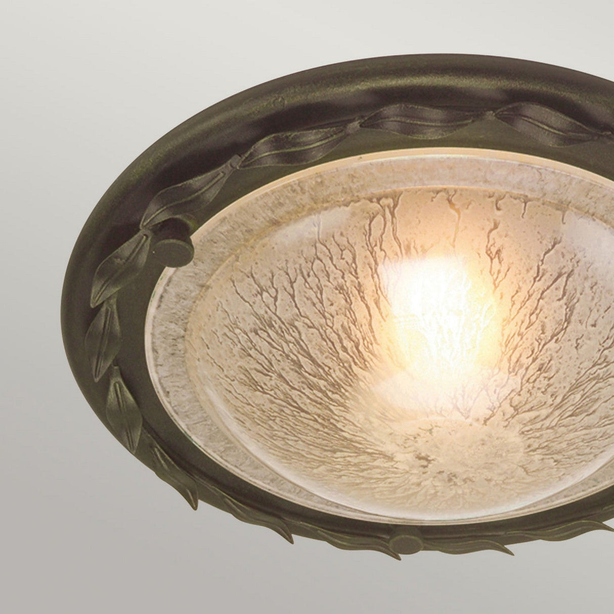 Close-up of the Olivia 1 Light Flush in Black/Gold Patina, a ceiling light fixture showcasing a decorative dark metal frame with leaf patterns. The glass shade diffuses warm, glowing light, providing a unique lighting solution with handcrafted elegance.