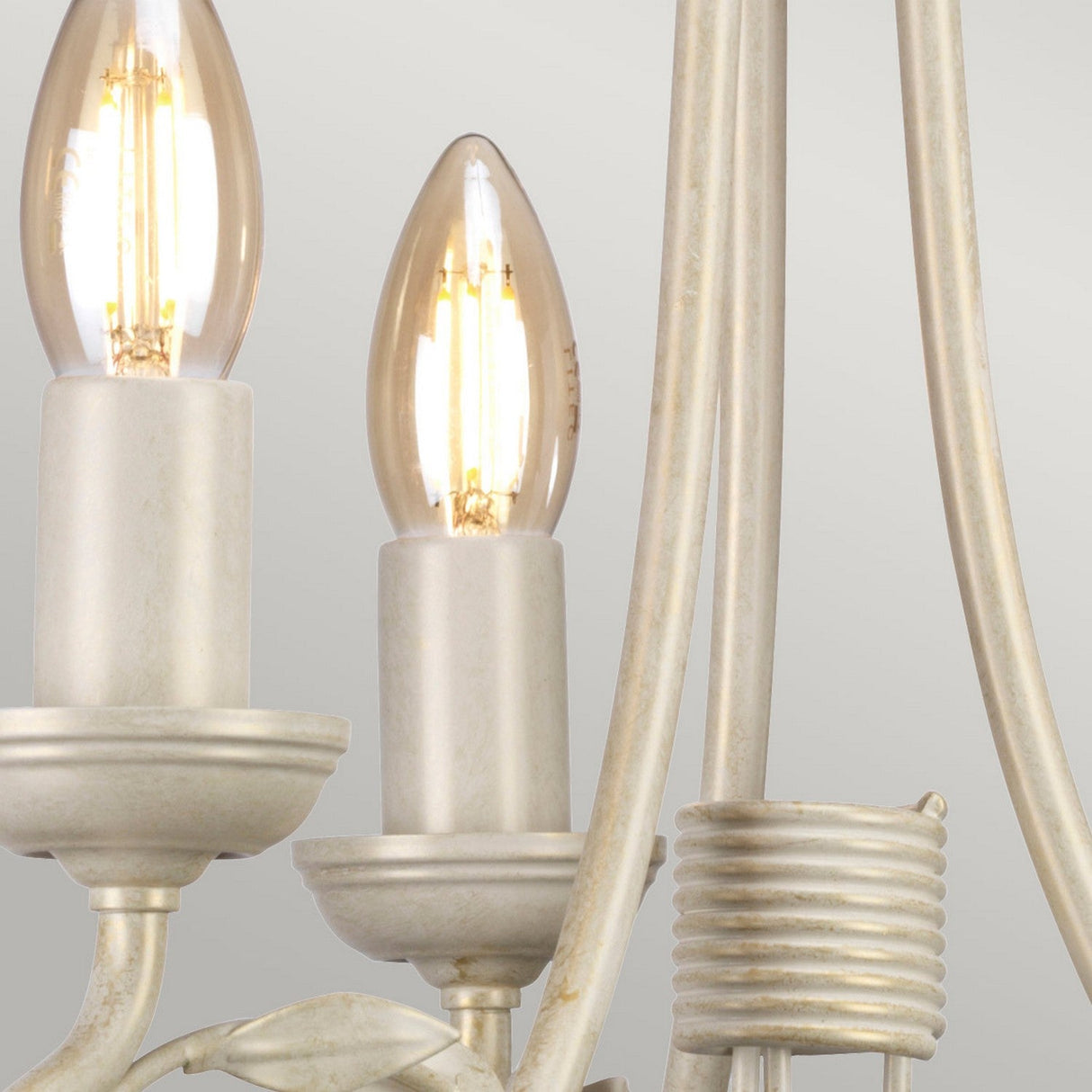 Close-up of the Olivia 3 Light Chandelier displaying its handmade vintage style with a pair of candle-like light bulbs emitting a soft glow. The fixture features an ivory/gold patina finish, gracefully curved metal rods, and a wrapped metal accent detail.