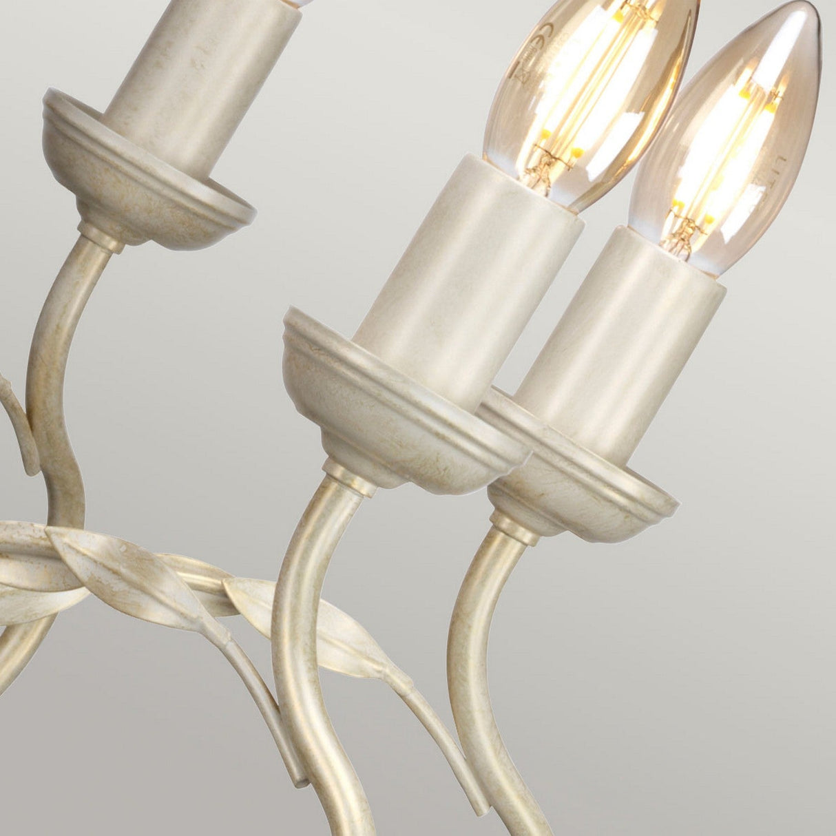 Detailed view of the Olivia 8 Light Chandelier in an ivory/gold patina finish, showcasing its elegant design. Each arm supports a vintage-style bulb that emits a warm glow against a neutral backdrop.