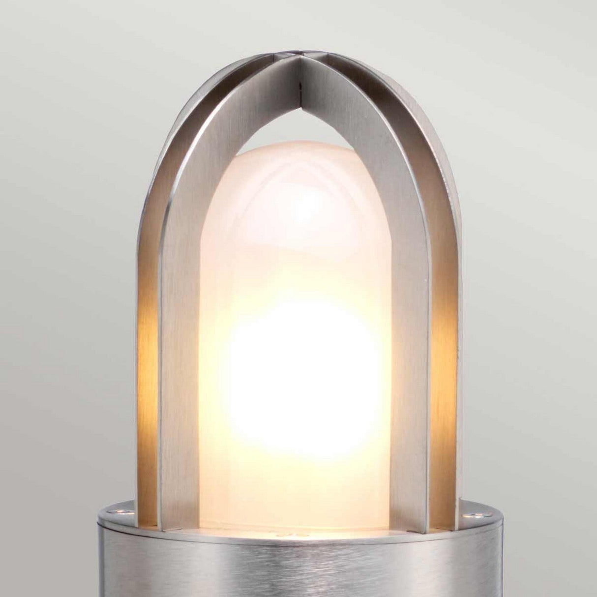 A contemporary and minimalist light fixture, the Paignton 1 Light Bollard - Stainless Steel showcases an arched metal frame enclosing a glowing, diffused bulb. The design incorporates sleek, brushed stainless steel and frosted glass to emit a soft, warm light against a neutral backdrop.