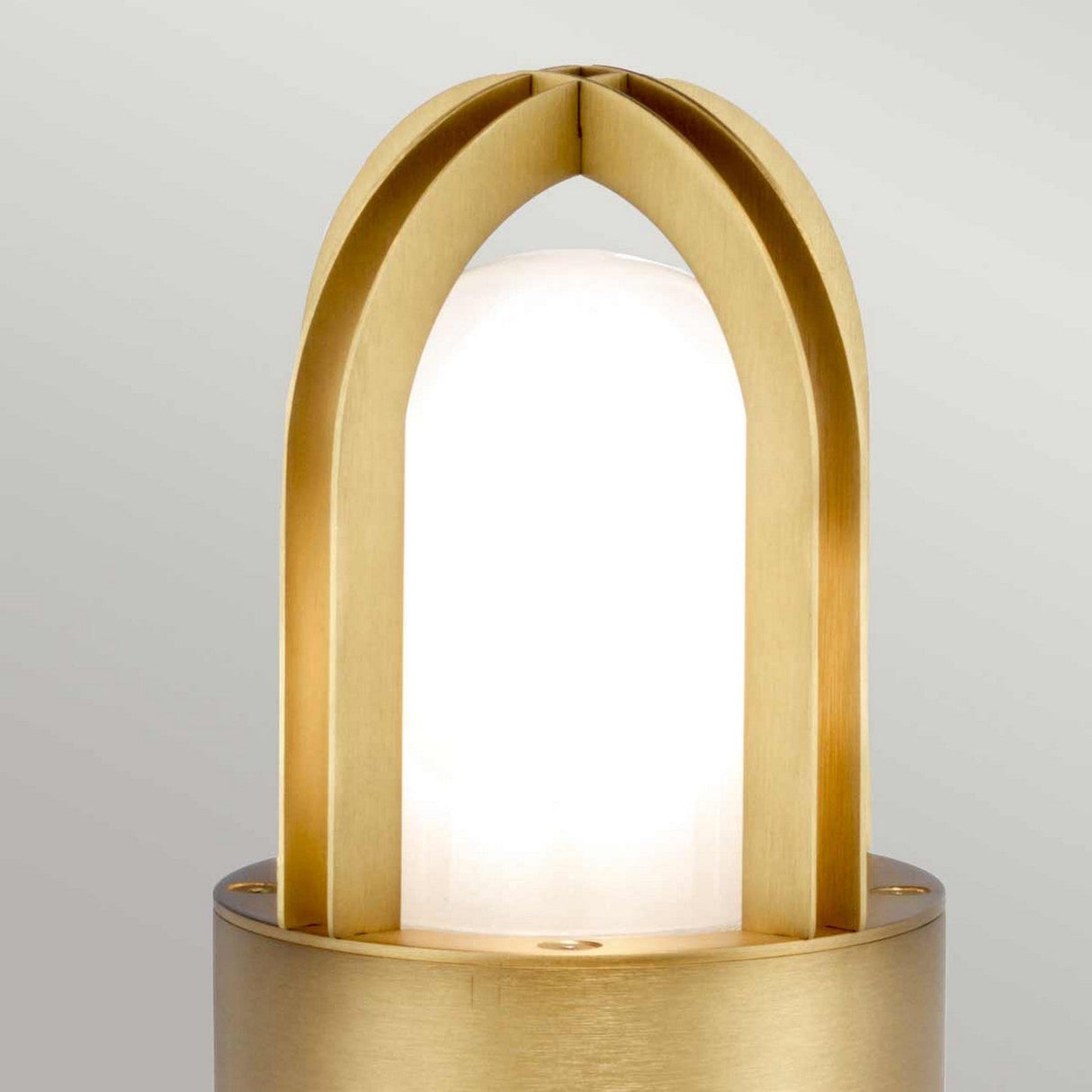 The Paignton 1 Light Mini Bollard in brushed brass features a contemporary gold arch design, encapsulating a frosted white bulb on a cylindrical base set against a neutral background.