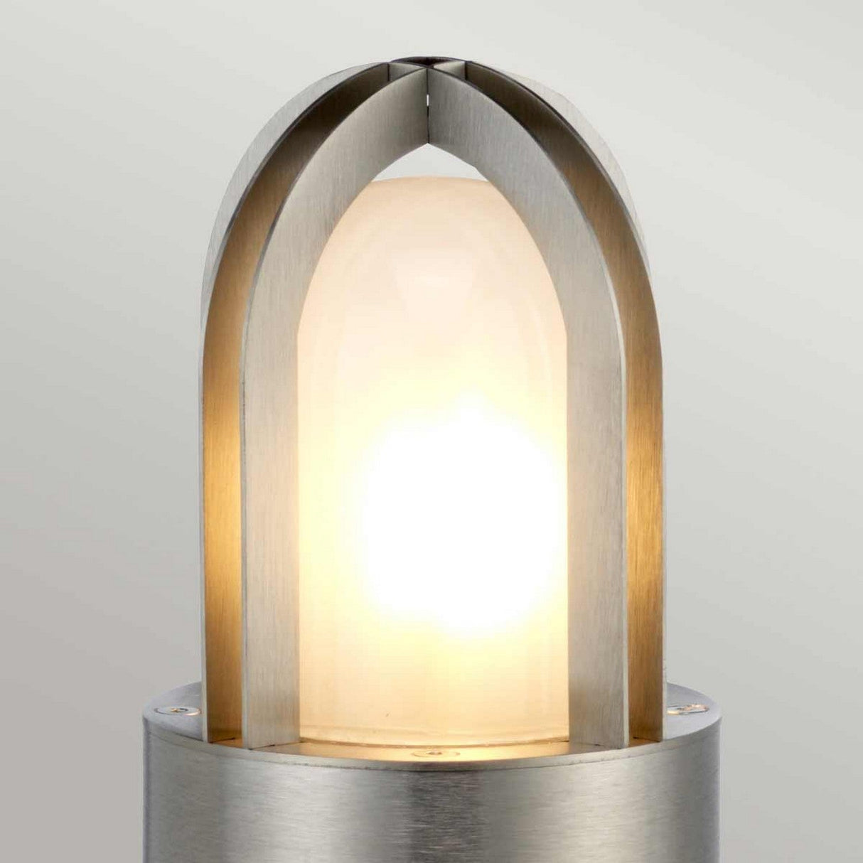 The Paignton 1 Light Mini Bollard in brushed stainless steel is a modern outdoor fixture with a cylindrical base. Its sleek design features an arch-shaped top that encloses a softly glowing bulb, casting warm light against a neutral backdrop.