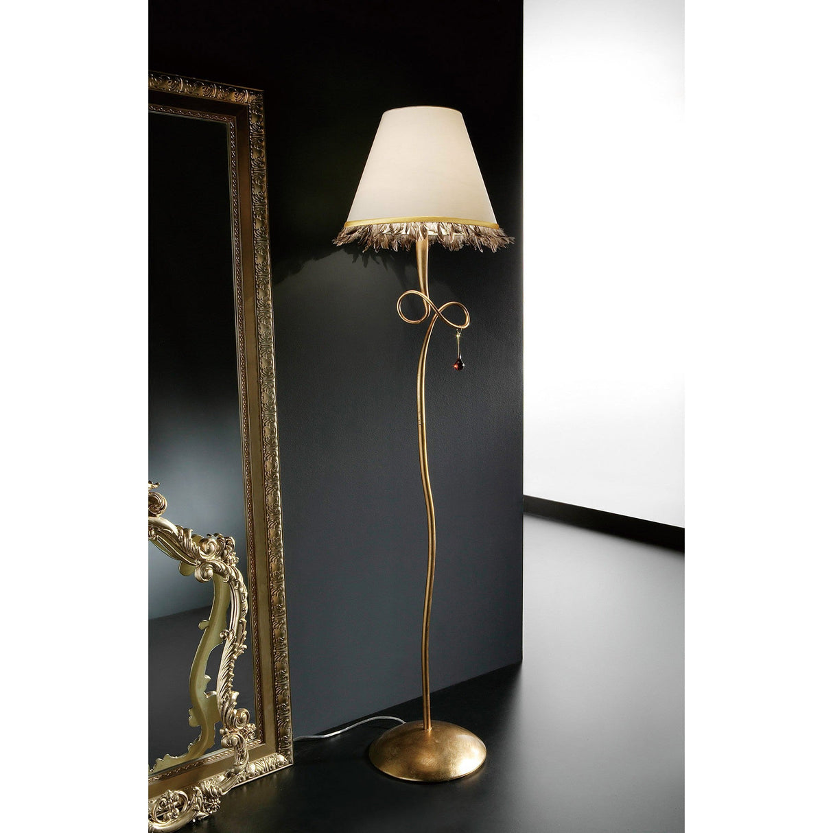 The Paola 1 Light Floor Lamp features a gold, curved stand with glass droplets and a cream shade, standing next to an ornate gold-framed mirror on a dark floor and wall with a bright white background.