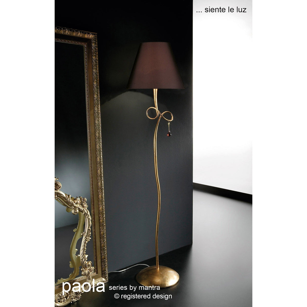 The Paola 1 Light Floor Lamp in gold features a curvy stand, cream shade, and shimmering glass droplets, standing beside a large ornate mirror and white wall. Text reads: paola series by mantra ... siente le luz © registered design.
