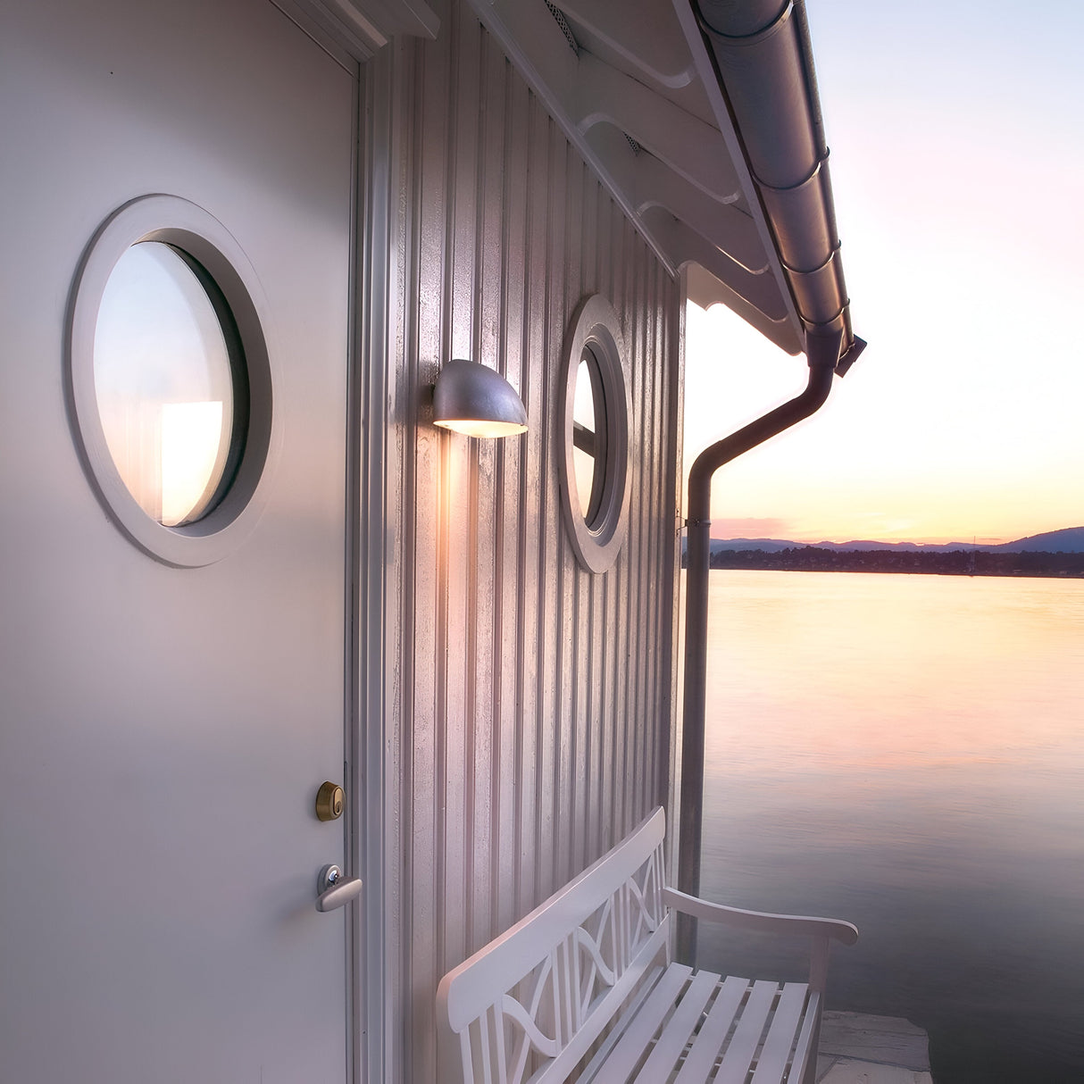 A tranquil lakeside scene includes a small white house with round windows and a bench outside. Beside the door, the Paris Outdoor Small 1 Light Wall Lantern in silver, with its IP43 rating, casts a warm glow over the calm water that reflects the soft pastel hues of sunset.