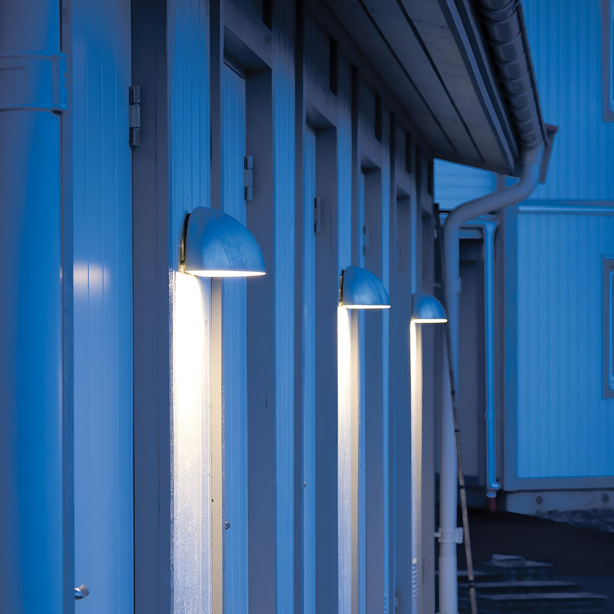A row of Paris Outdoor Large 1 Light Wall Lanterns - Silver casts a soft white glow on the vertical panels of a building exterior at dusk. The lanterns, with their blue tint, illuminate the structure, which features multiple doors, gutters, and a sloped roof. The scene exudes calm and tranquility.