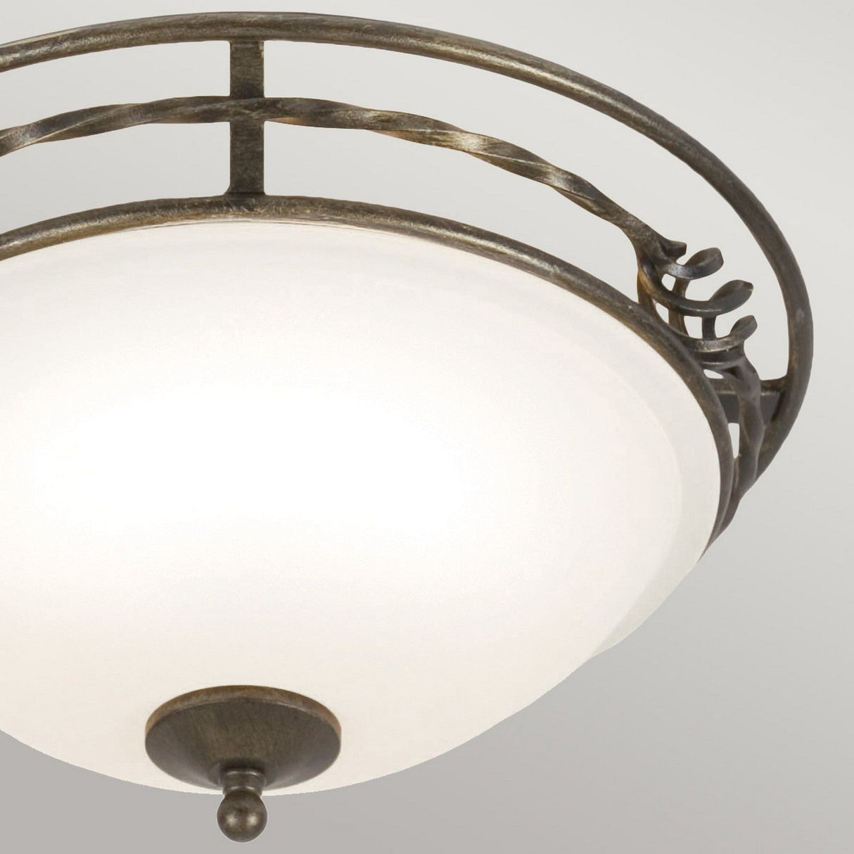 A close-up of the ceiling-mounted Pembroke 2 Light Flush Light - Black/Gold, showcasing its frosted glass shade. It boasts a hand-forged iron frame with intricate scrollwork detailing and a small decorative finial at the bottom. The background is a light grey color.