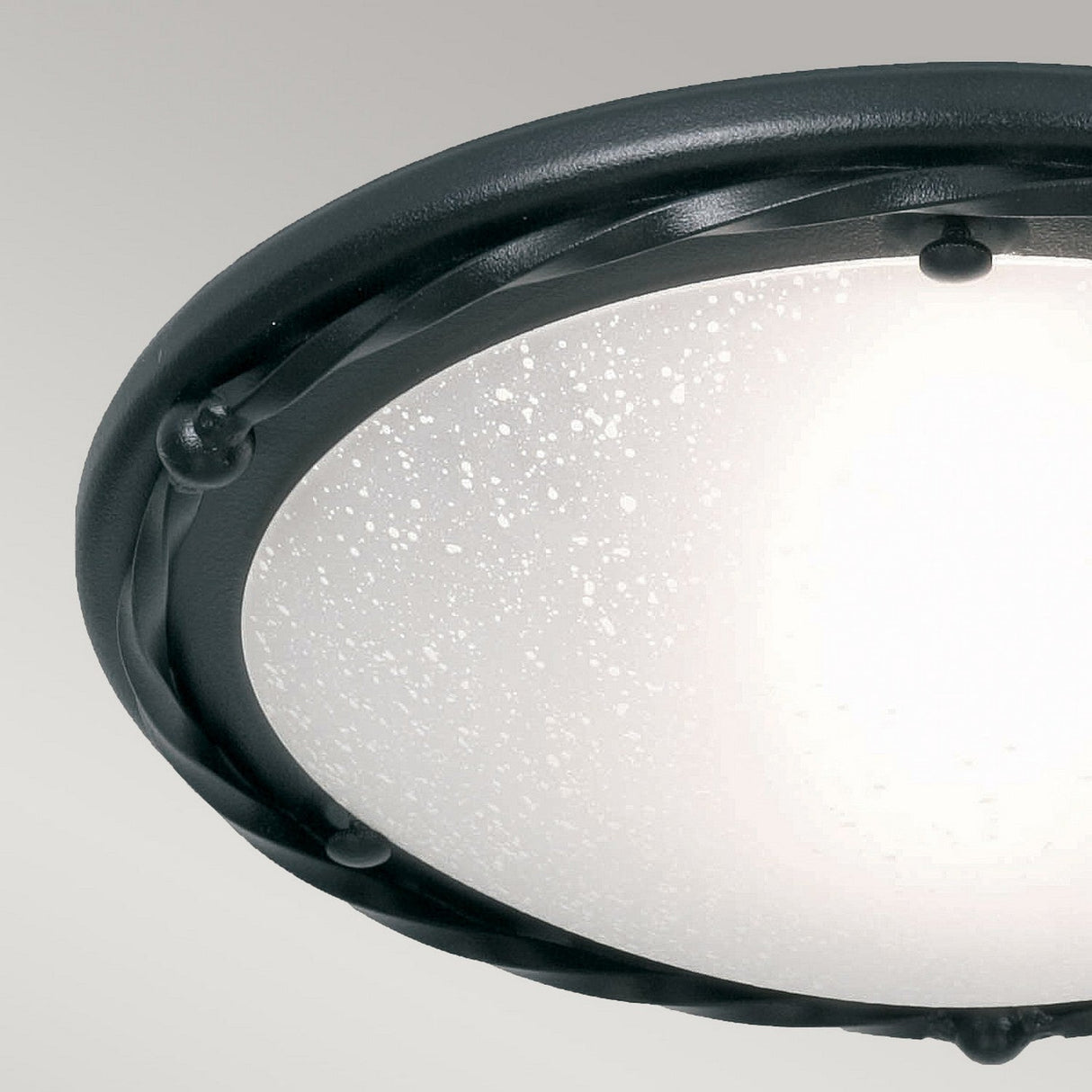 A close-up of the Pembroke 1 Light Flush Light - Black showcases modern elegance with its circular frosted glass shade and hand-forged iron rim. The glass is adorned with delicate speckled patterns, and the fixture is seamlessly mounted on a light-colored ceiling.