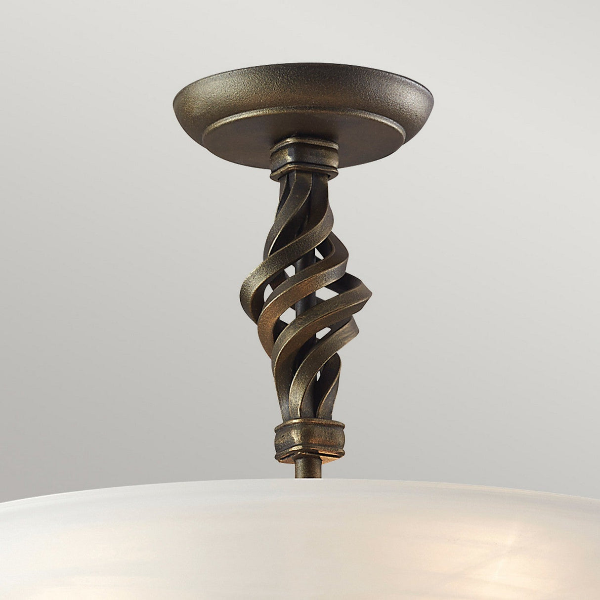 A close-up of the Pembroke 3 Light Semi Flush fixture reveals a beautifully crafted, intricately twisted hand-forged iron design with a bronze finish that supports a frosted glass shade. It is mounted against a light gray ceiling background.