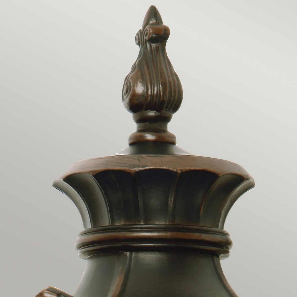Close-up of an ornamental finial on an old bronze metal object, possibly a decorative lidded jar or vase. The finial features a carved, ornate design reminiscent of a stylized flame or leaf pattern, similar to the intricate detailing seen on the Philadelphia 3 Light Outdoor Large Wall Light in Old Bronze.