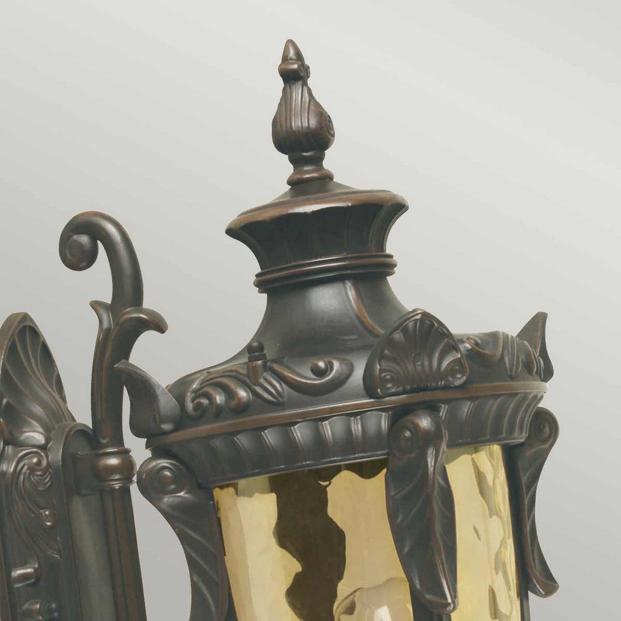 Close-up of the Philadelphia 1 Light Outdoor Medium Wall Light in Old Bronze, featuring a dark metal frame and a curved top. Exuding traditional elegance, it showcases decorative leaf patterns and scroll elements, complemented by yellowish glass panels. Ideal for outdoor lighting, it is set against a plain light gray backdrop.