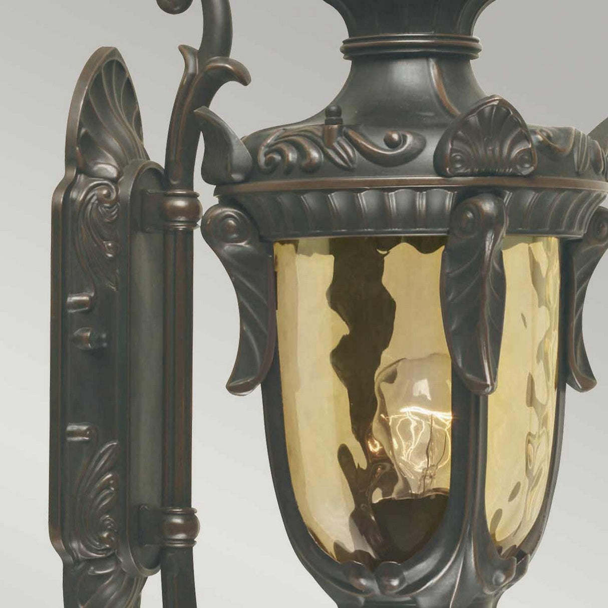 The Philadelphia 1 Light Outdoor Medium Wall Light - Old Bronze showcases traditional elegance with its intricate detailing and warm yellow glass casing enclosing a light bulb, set against a plain gray background.