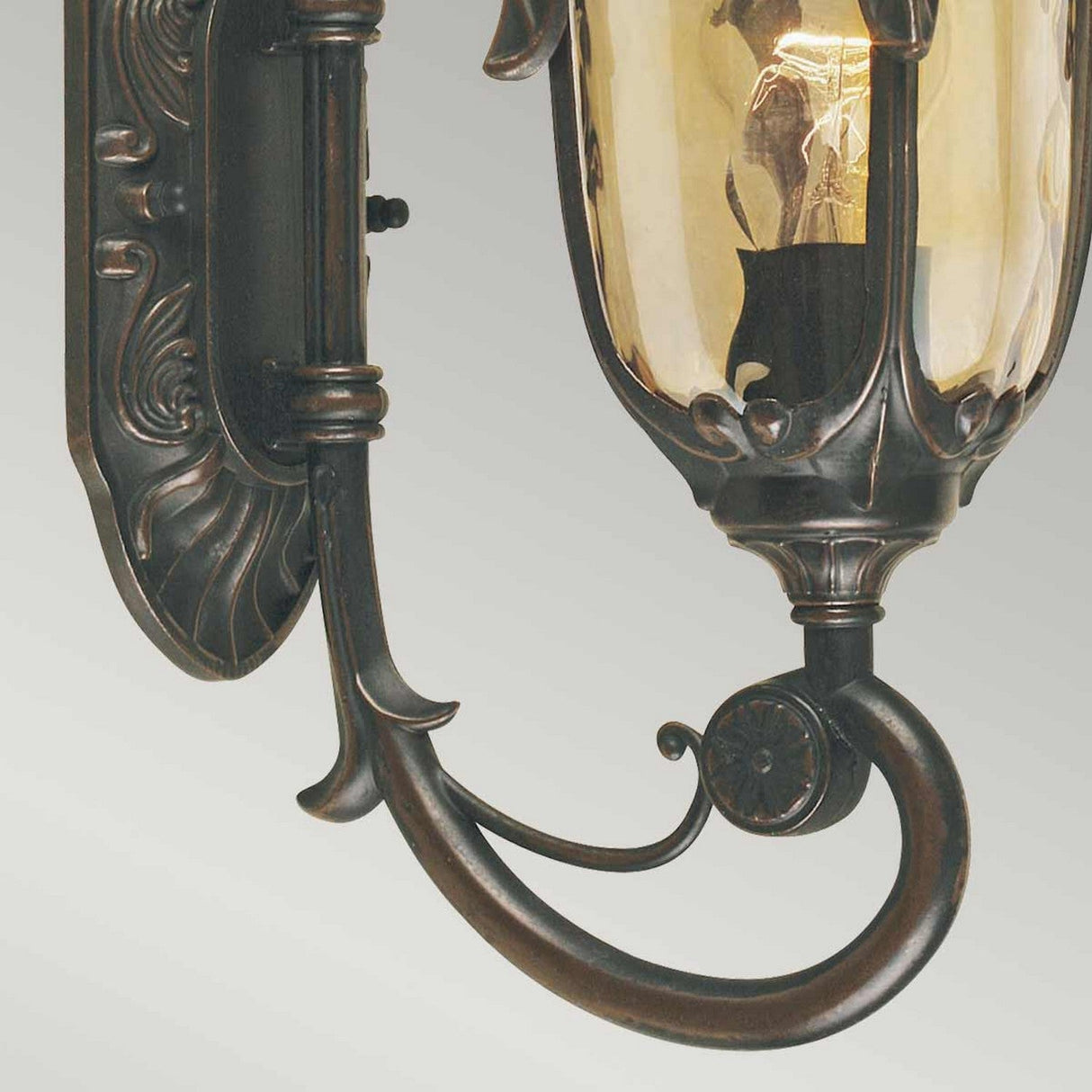 The Philadelphia 1 Light Outdoor Small Wall Light in Old Bronze features a period-inspired design with an ornate bronze finish and a clear, amber-tinted glass shade. The exposed bulb enhances the old bronze patina, and the elegant curved detailing stands out against the light gray background.